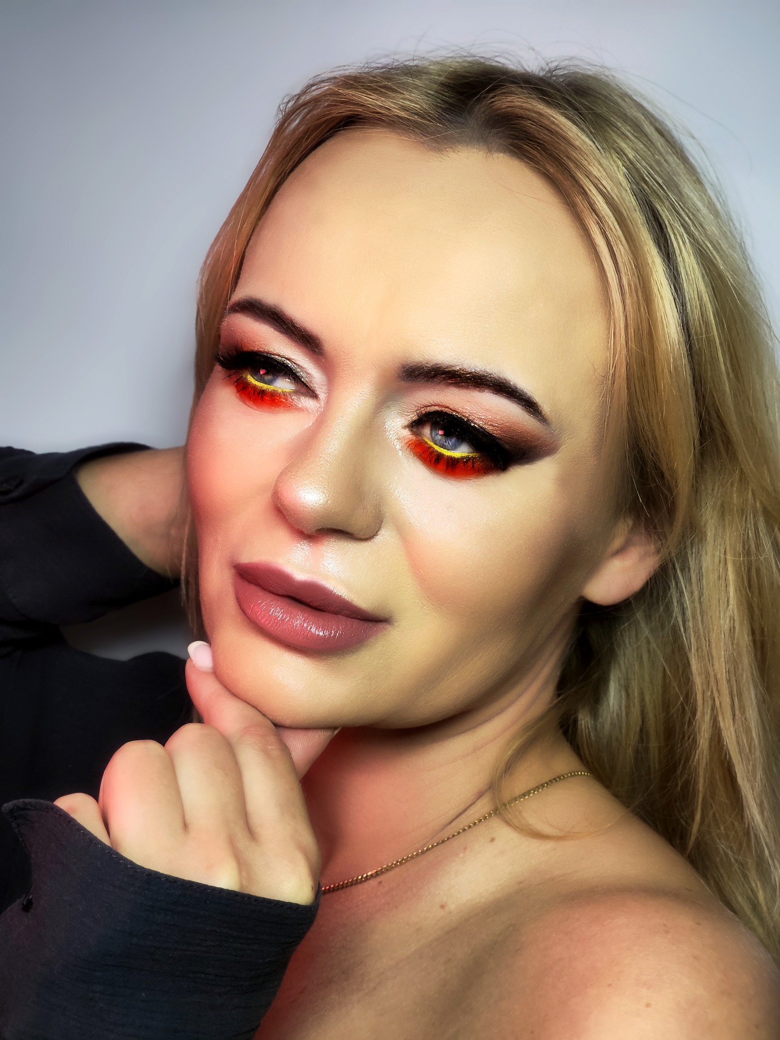 Portret kobiety z intensywnym, artystycznym zdobieniem powiek w odcieniach czerwieni, żółci i czerni; usta w kolorze nude, delikatna złota biżuteria na szyi.