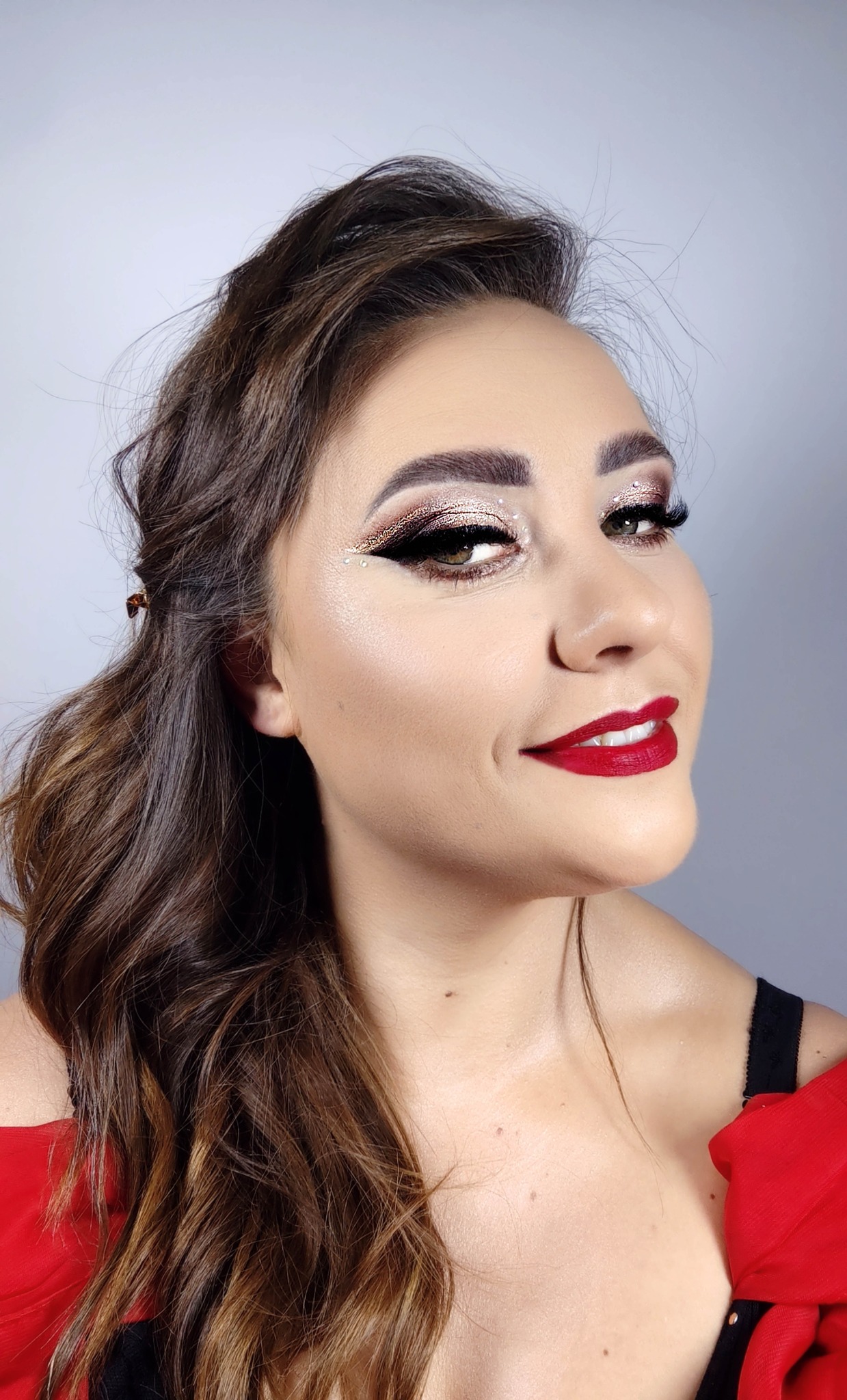 Portret kobiety z błyszczącym cieniem na powiekach, sztucznymi rzęsami i intensywnie czerwoną szminką, uśmiechająca się lekko w stronę widza.