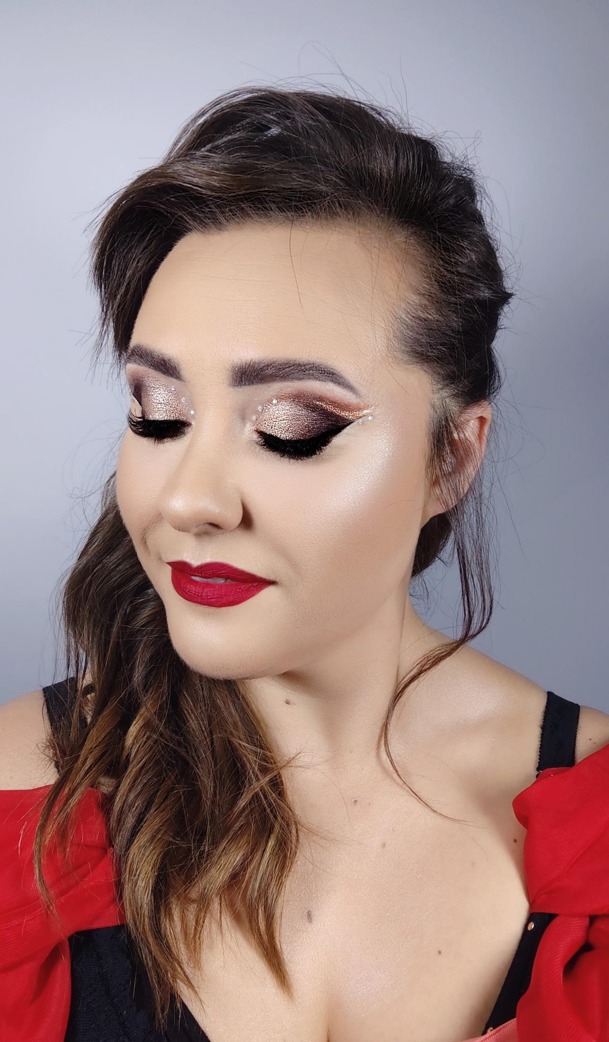 Portret kobiety z zamkniętymi oczami, prezentującej wieczorowy look z brokatowymi cieniami, czarnym eyelinerem i błyszczącymi kamyczkami przy zewnętrznym kąciku oka oraz intensywnie czerwoną szminką.