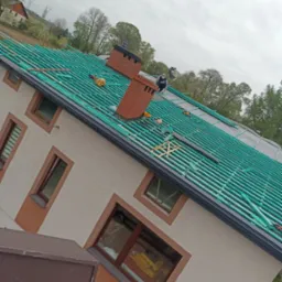 Montaż łat na dachu domu jednorodzinnego z ceglanym kominem; widoczny pracownik w trakcie pracy, narzędzia i materiały budowlane.