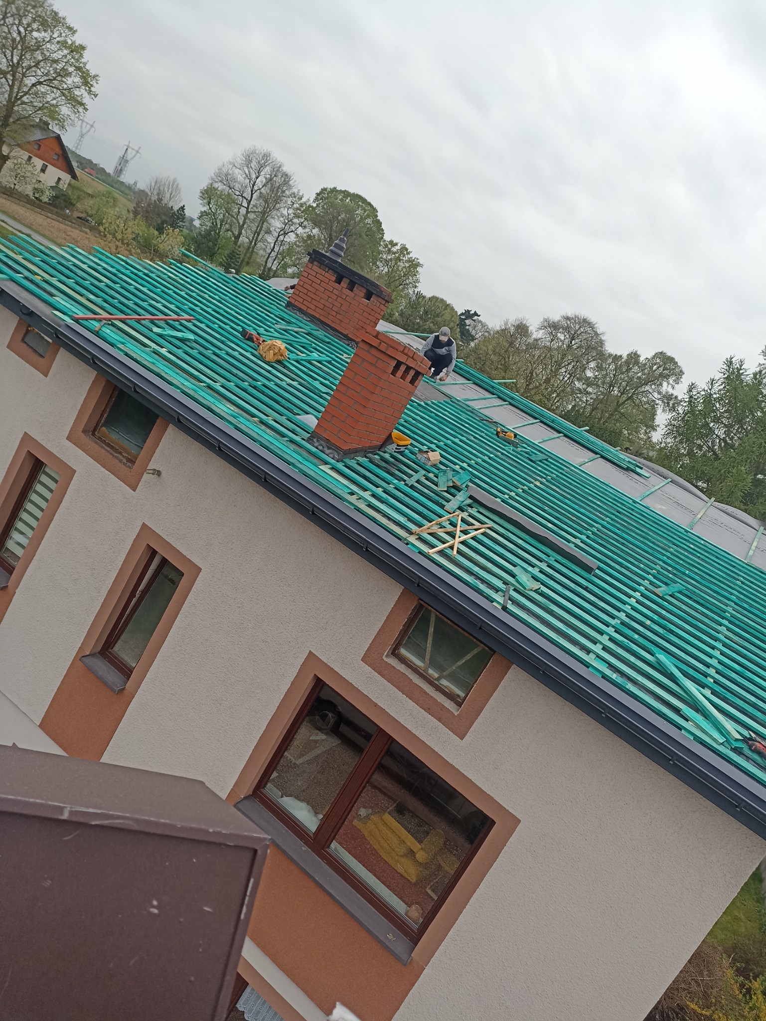 Montaż łat na dachu domu jednorodzinnego z ceglanym kominem; widoczny pracownik w trakcie pracy, narzędzia i materiały budowlane.