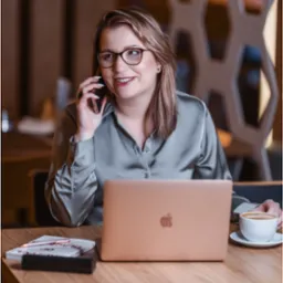 Kobieta w okularach rozmawia przez telefon, siedząc przy drewnianym stole z laptopem, książką, długopisem i filiżanką kawy; w tle dekoracyjna ściana.