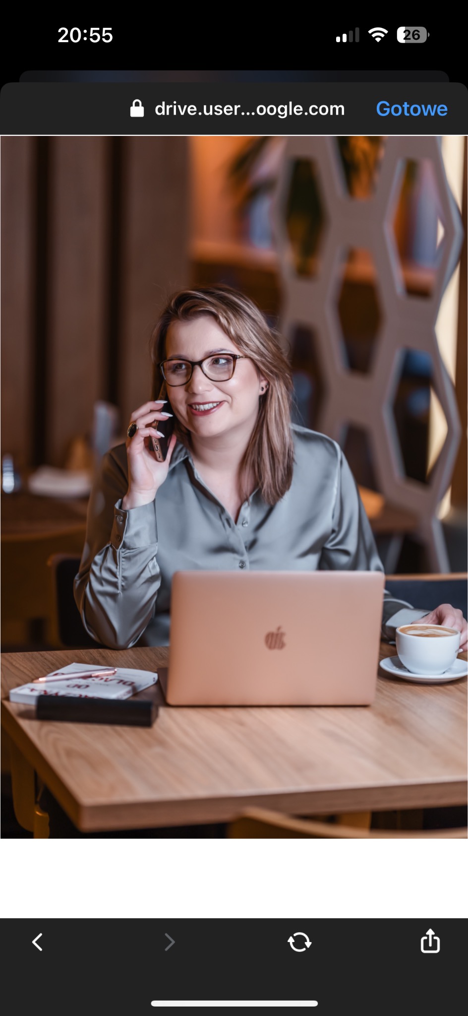 Kobieta w okularach rozmawia przez telefon, siedząc przy drewnianym stole z laptopem, książką, długopisem i filiżanką kawy; w tle dekoracyjna ściana.