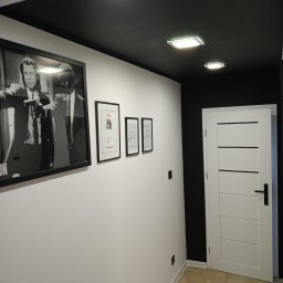 Filip Zawadzki - Korytarz po remoncie w Olecku z czarnym sufitem, białymi ścianami ozdobionymi ramkami i białymi drzwiami z czarną klamką.