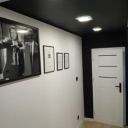 Korytarz po remoncie w Olecku z czarnym sufitem, białymi ścianami ozdobionymi ramkami i białymi drzwiami z czarną klamką.