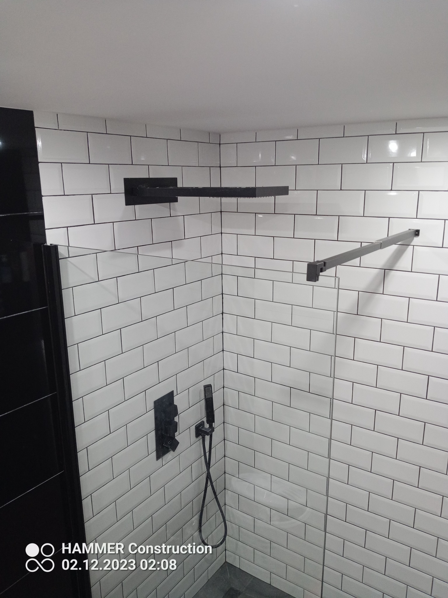 Nowoczesna kabina prysznicowa z białymi, prostokątnymi płytkami ułożonymi w cegiełkę, czarna armatura i szklane drzwi, minimalistyczny design.