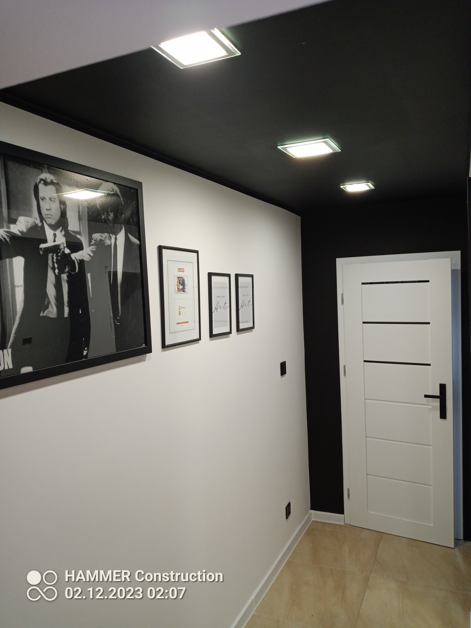 Korytarz po remoncie w Olecku z czarnym sufitem, białymi ścianami ozdobionymi ramkami i białymi drzwiami z czarną klamką.