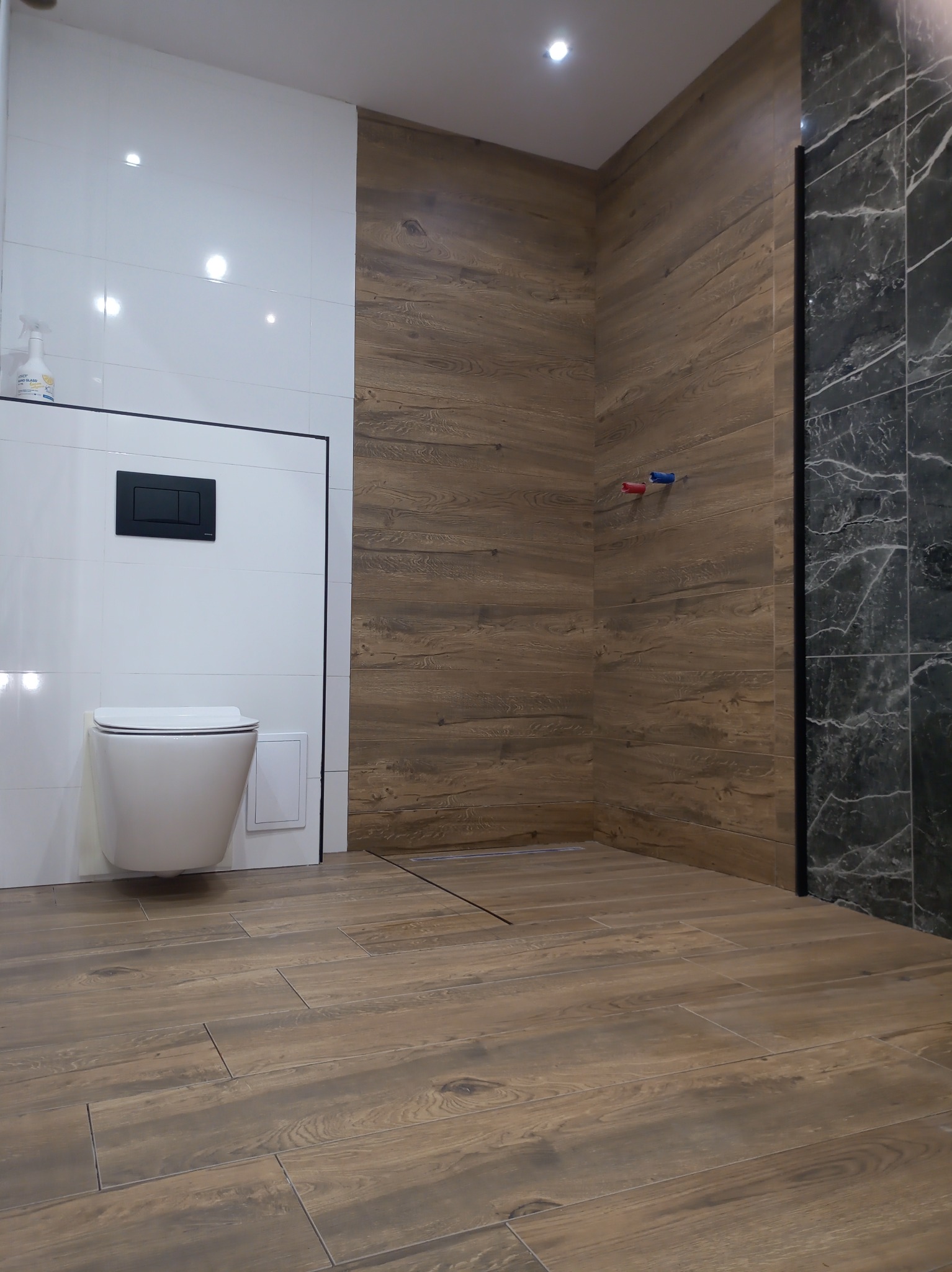 Nowoczesna łazienka z wiszącą toaletą, białymi płytkami, drewnopodobnymi panelami ściennymi i podłogowymi oraz czarnymi akcentami, w tym odpływem liniowym i marmurową ścianą prysznicową.