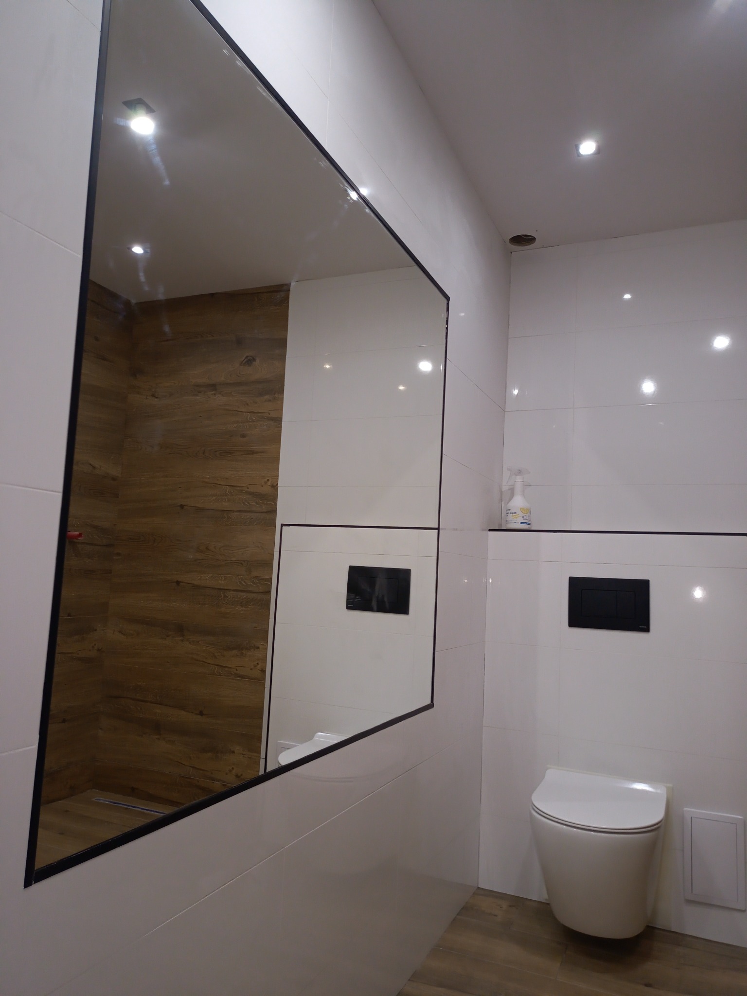 Nowoczesna łazienka z białymi płytkami, dużym lustrem z czarną ramą, podwieszaną toaletą i drewnopodobną podłogą, widok na kabinę prysznicową w odbiciu lustra.