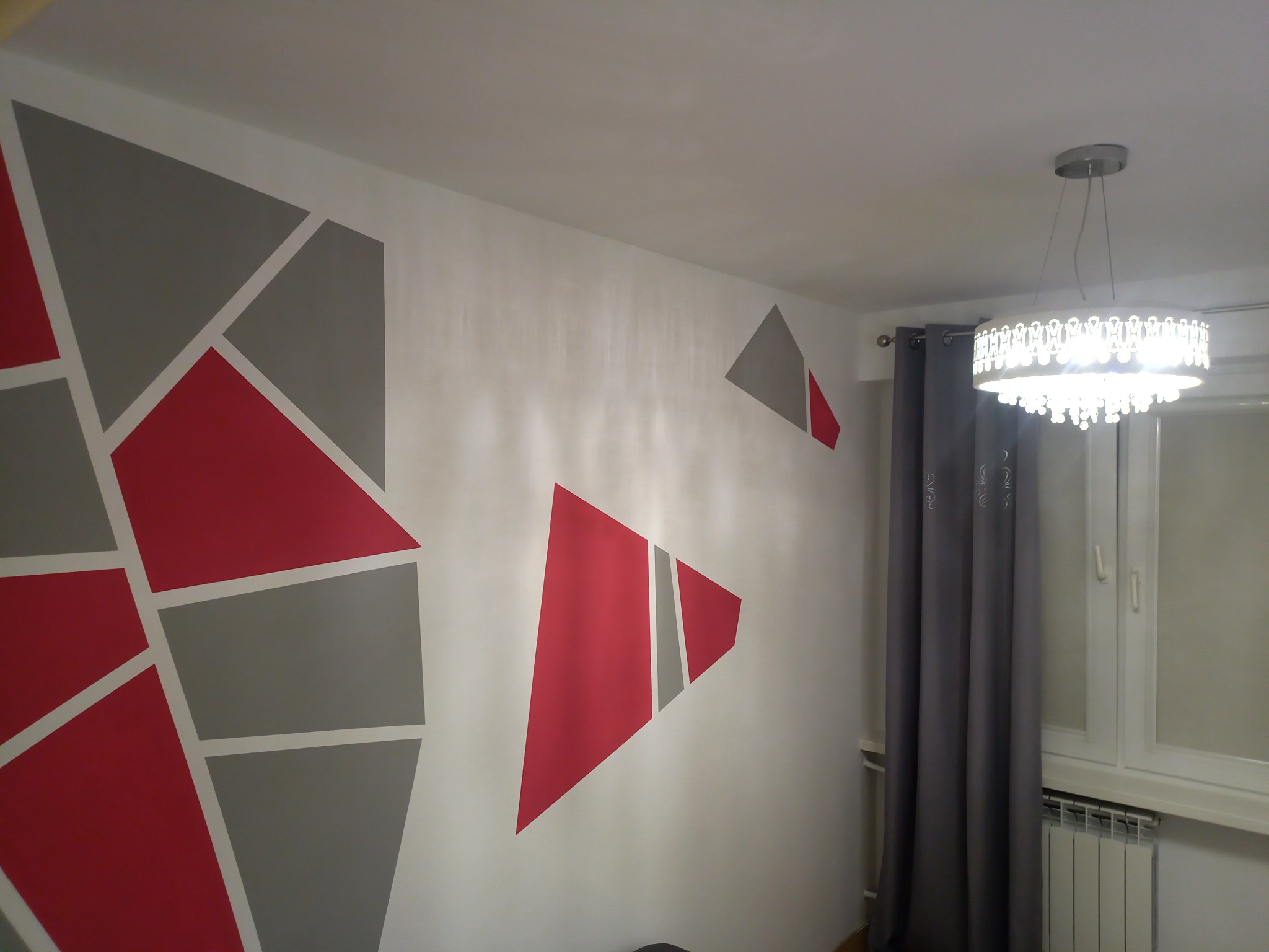 Ściana z geometrycznym wzorem w odcieniach czerwieni, szarości i bieli, oświetlona przez nowoczesny, okrągły żyrandol z kryształkami; obok okno z szarymi zasłonami.