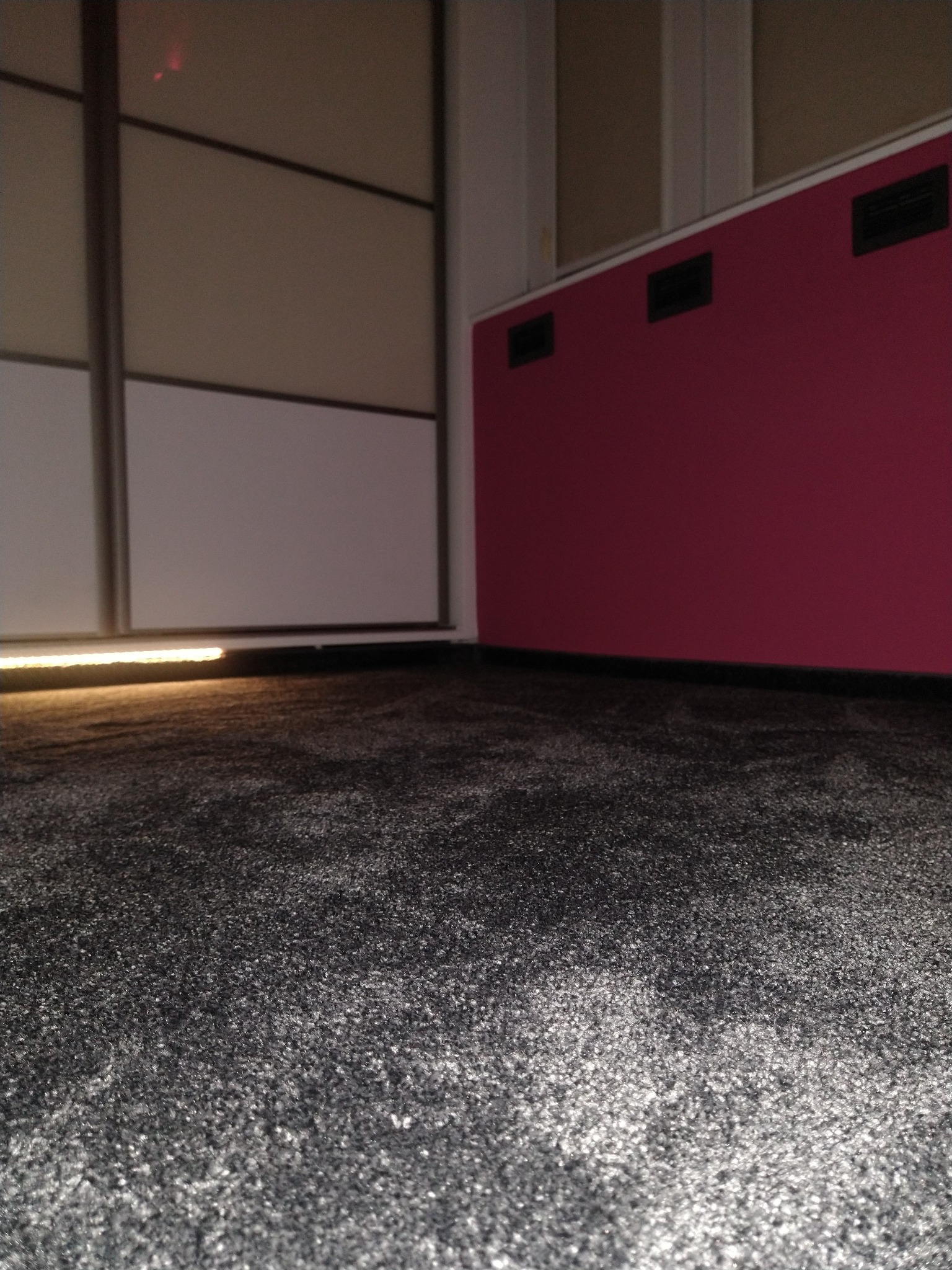 Szary dywan z krótkim włosiem w rogu pokoju, widoczna dolna część przesuwnej szafy z oświetleniem LED i fragment bordowej ściany z kratkami wentylacyjnymi.