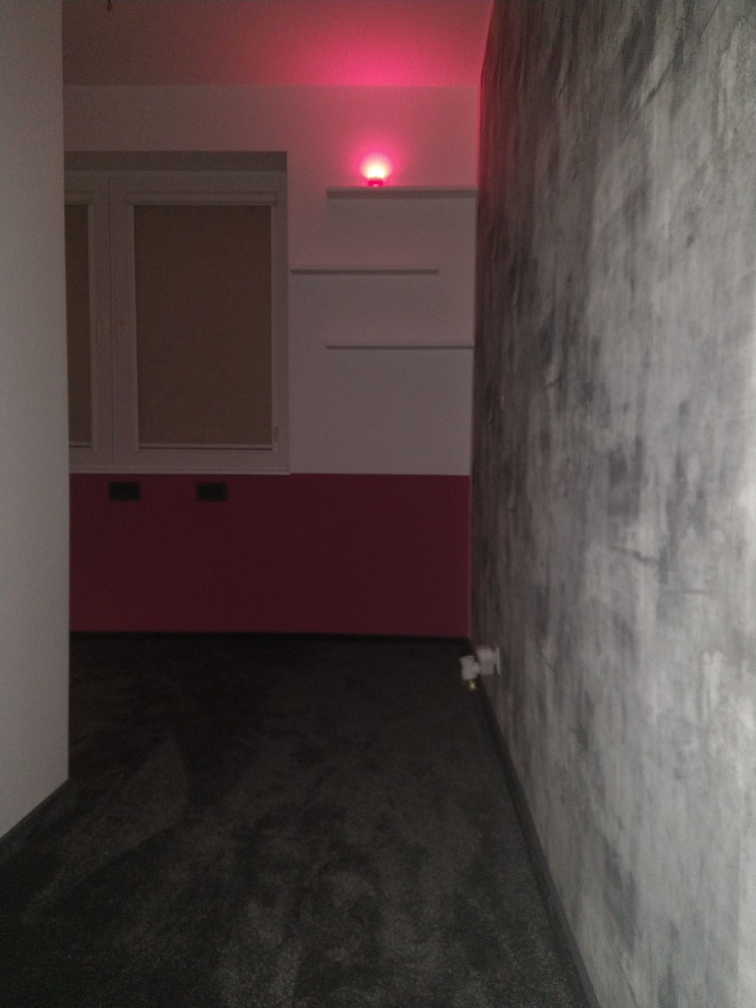 Pokój po remoncie z szarą ścianą o fakturze betonu, czerwonym pasem na ścianie z oknem i czarną podłogą, oświetlony czerwonym światłem z góry.