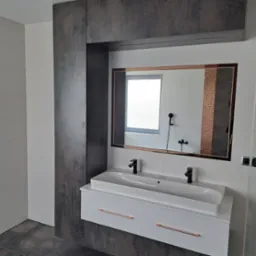 Nowoczesna łazienka z podwójną białą umywalką nablatową, czarną armaturą i szafką z miedzianymi uchwytami, wbudowana w ciemną zabudowę meblową z lustrem w miedzianej ramie, odbijającym prysznic.