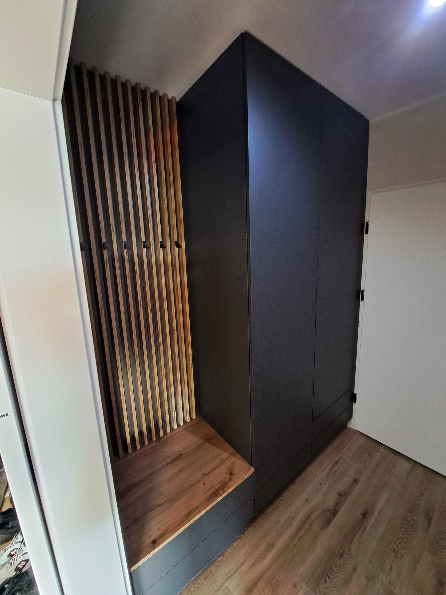 Ciemnoszara szafa wnękowa z drewnianymi listwami i siedziskiem w przedpokoju, widoczne haki na ubrania.