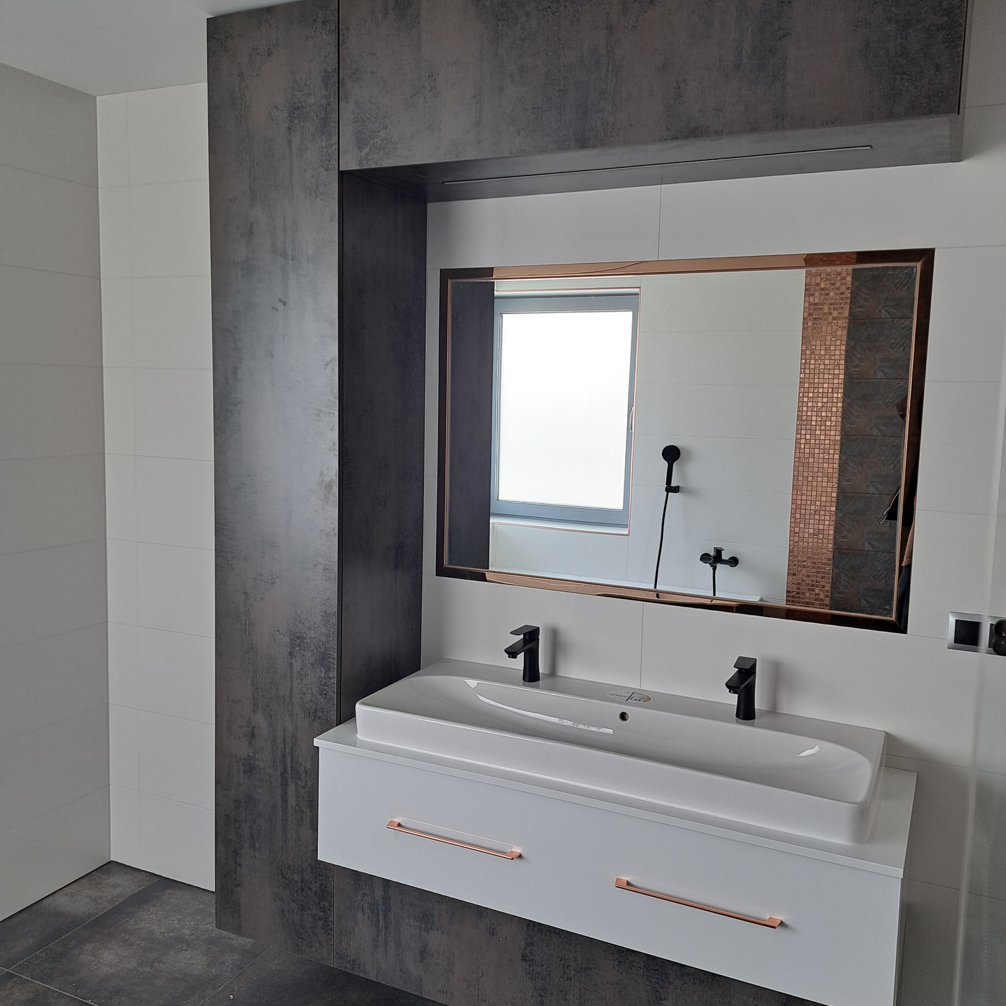 Nowoczesna łazienka z podwójną białą umywalką nablatową, czarną armaturą i szafką z miedzianymi uchwytami, wbudowana w ciemną zabudowę meblową z lustrem w miedzianej ramie, odbijającym prysznic.