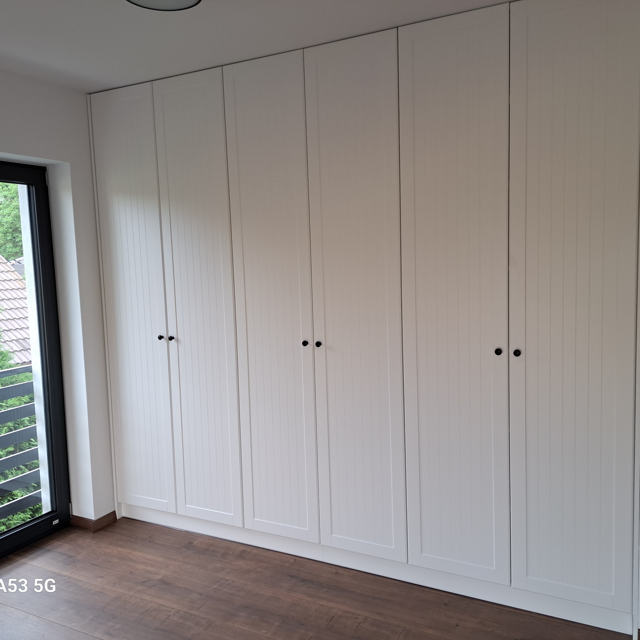 Biała, zabudowana szafa wnękowa z pionowymi frezowaniami na frontach drzwi, z czarnymi, okrągłymi uchwytami, na tle drewnianej podłogi i okna z widokiem na zieleń.