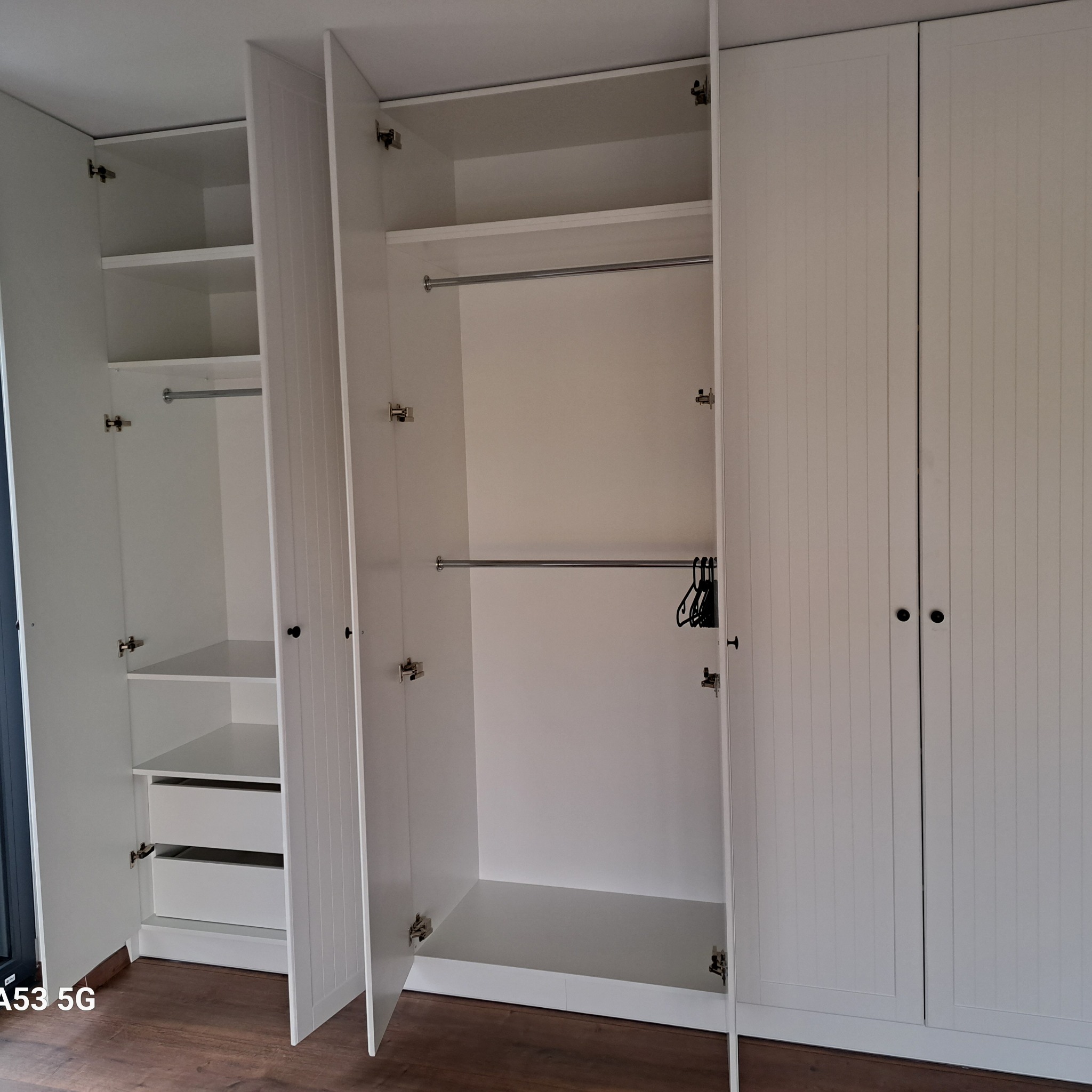 Biała szafa wnękowa z otwartymi drzwiami, widoczne półki, szuflady i drążek na wieszaki, minimalistyczny design.