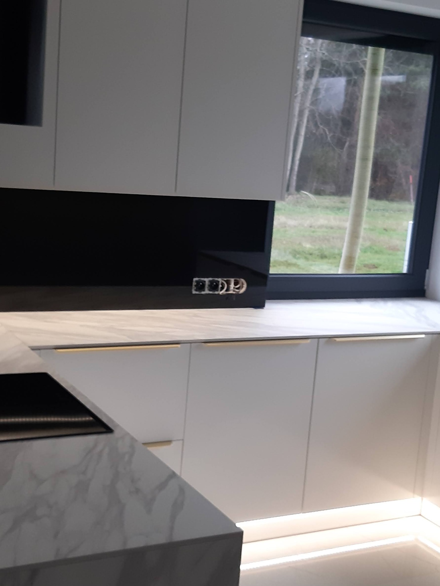 Biała kuchnia z marmurowym blatem, złotymi uchwytami i czarnym panelem nad blatem. Widoczne gniazdka elektryczne i okno z widokiem na drzewa.