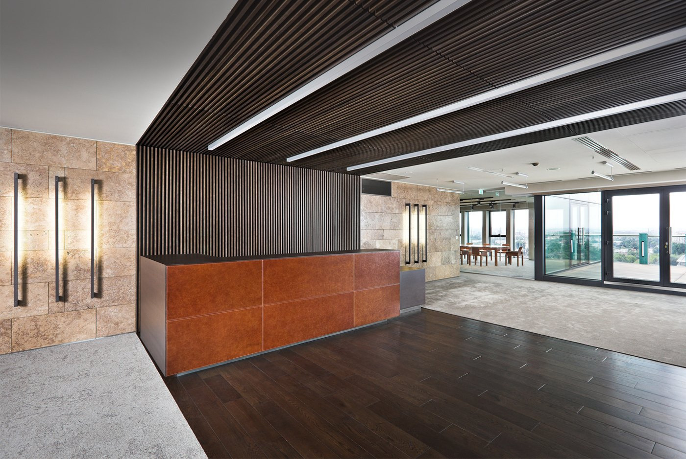 Recepcja biurowa z brązową ladą, drewnianym sufitem lamelowym i kamienną ścianą z podłużnymi lampami.