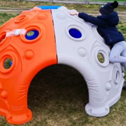 Dziecko wspina się na dwukolorową (pomarańczowo-szarą) kopułę do wspinaczki z otworami, umieszczoną na trawie. Na pomarańczowej części widoczna taśma klejąca z zabezpieczeniem.