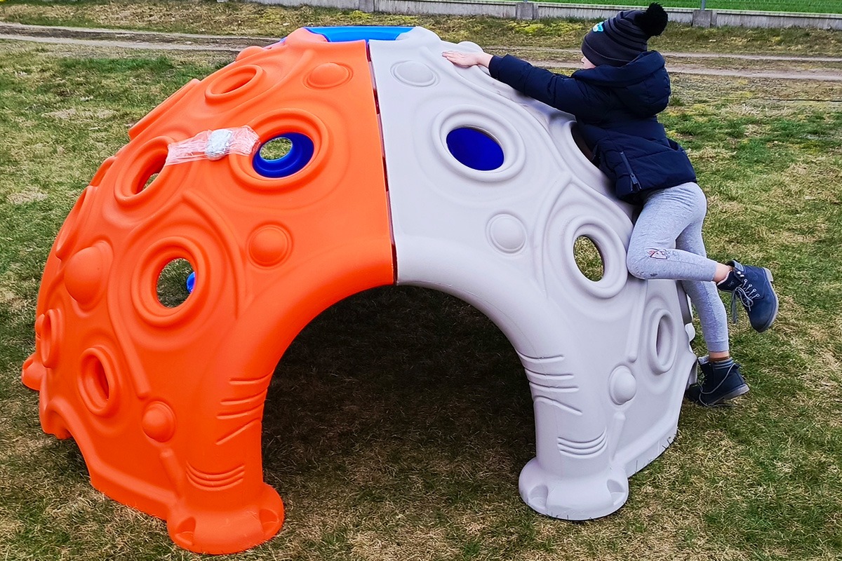 Dziecko wspina się na dwukolorową (pomarańczowo-szarą) kopułę do wspinaczki z otworami, umieszczoną na trawie. Na pomarańczowej części widoczna taśma klejąca z zabezpieczeniem.