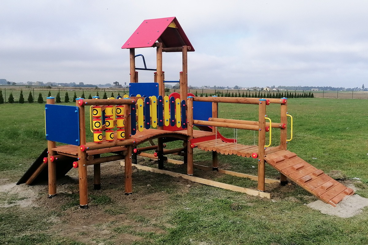 Kolorowy, drewniany plac zabaw z wieżą, zjeżdżalnią i drabinkami na trawiastym terenie, widok z przodu, w tle rząd drzew.