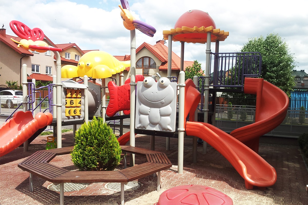 Kolorowy plac zabaw z motywami zwierzęcymi, otoczony sześciokątną ławką i małym drzewkiem iglastym, na tle domów jednorodzinnych.