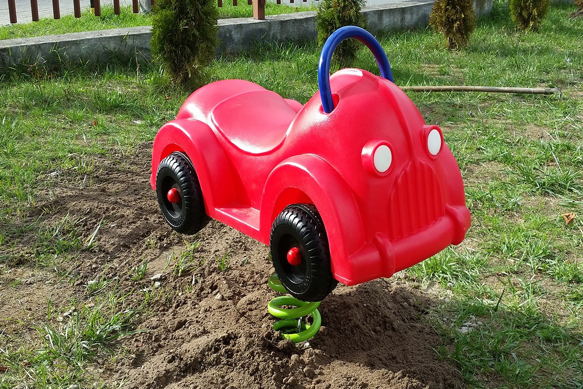 Czerwony, plastikowy samochód na sprężynie, element placu zabaw, z niebieskim uchwytem, osadzony w ziemi z trawą.