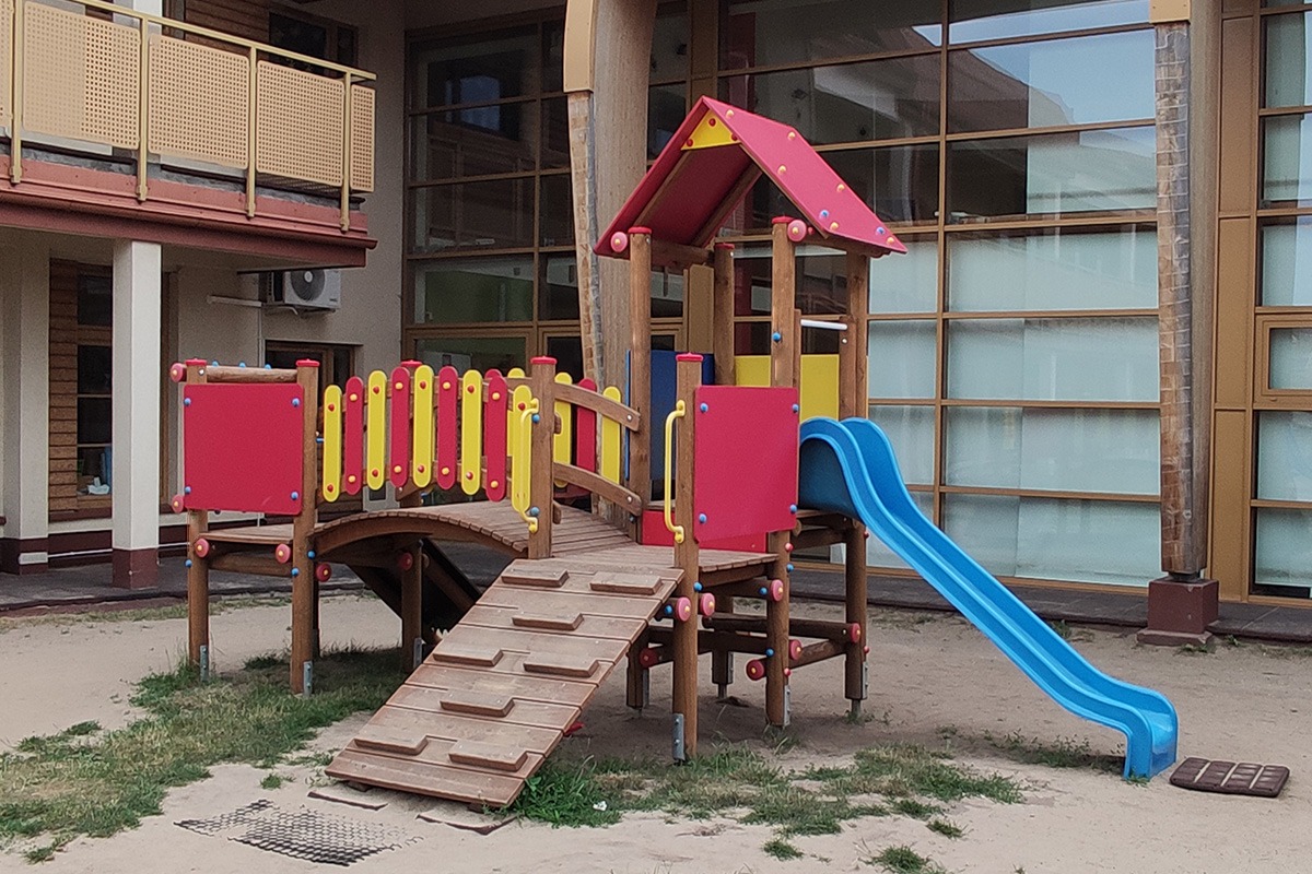 Kolorowy plac zabaw z drewnianym mostkiem, zjeżdżalnią i schodami na tle budynku z dużymi oknami.