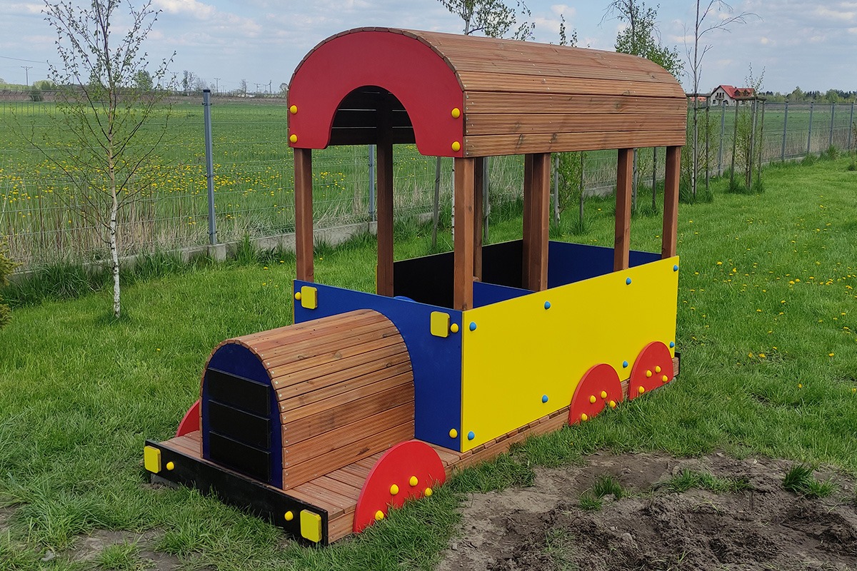 Kolorowa drewniana lokomotywa dla dzieci na trawniku z widokiem na pole i ogrodzenie.