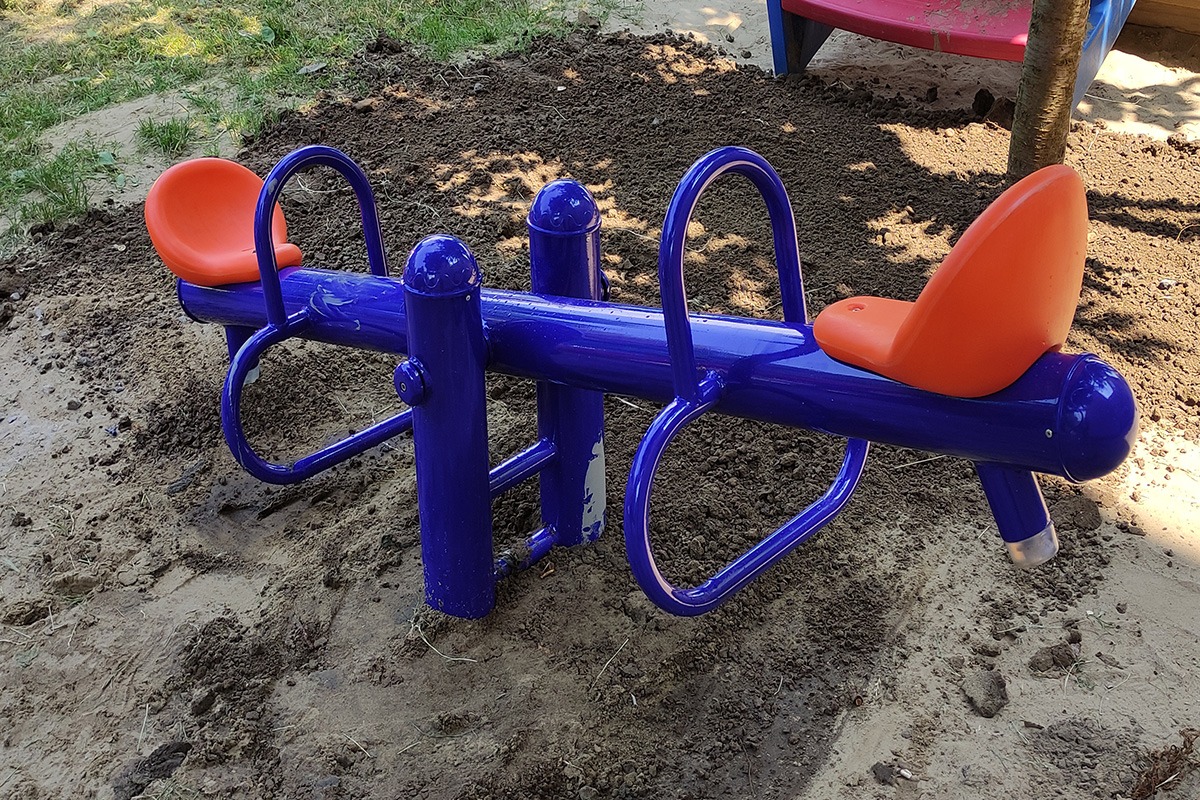 Niebieska huśtawka wahadłowa z pomarańczowymi siedziskami na placu zabaw, otoczona świeżo usypaną ziemią.