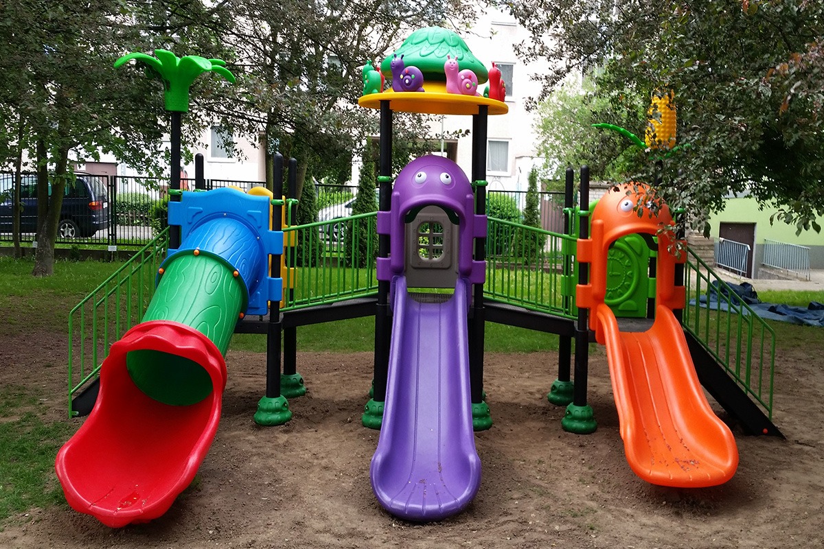 Kolorowy zestaw zabawowy z trzema zjeżdżalniami o różnych kształtach i kolorach: tunel w barwach drewna, fioletowa zjeżdżalnia z motywem twarzy i pomarańczowa zjeżdżalnia z motywem potwora, na tle...
