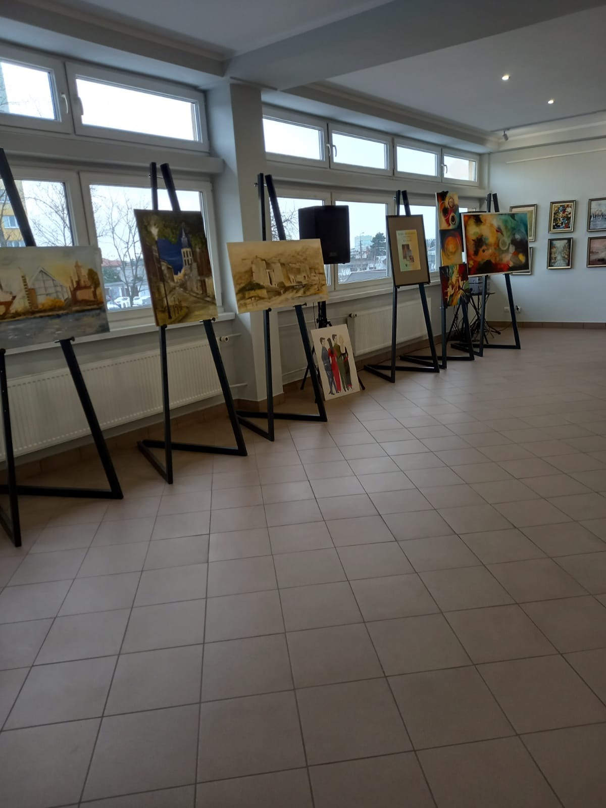 Wystawa obrazów na sztalugach w jasnym pomieszczeniu galerii we Włocławku, z widokiem na miasto przez okna w tle.