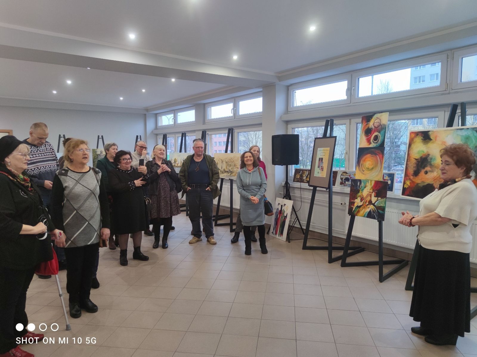 Wernisaż obrazów: grupa osób ogląda wystawę prac malarskich na sztalugach w jasnym pomieszczeniu z oknami.