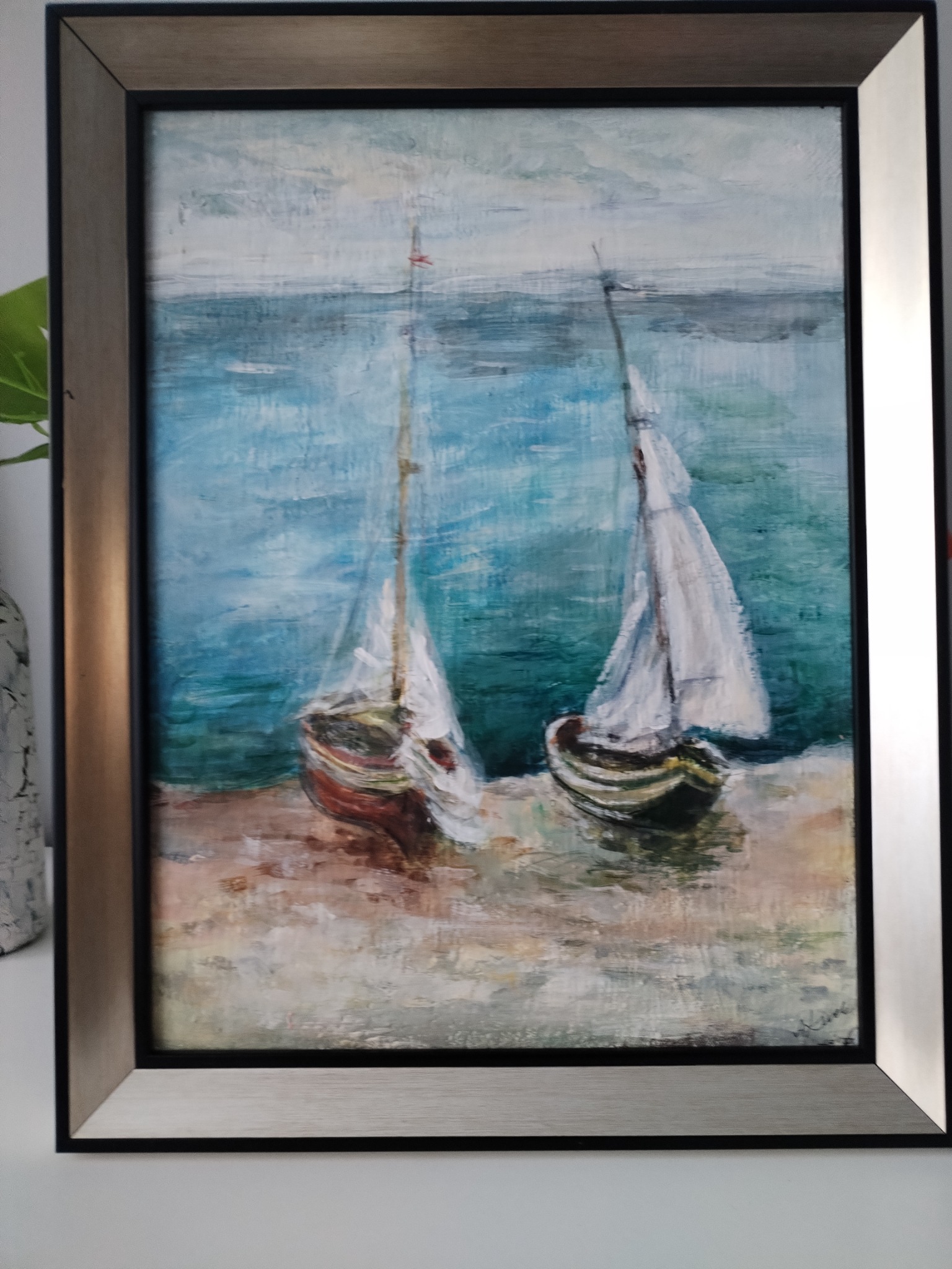 Oprawiony obraz olejny przedstawiający dwie łodzie żaglowe na brzegu morza, w spokojnych odcieniach błękitu i beżu, z widocznymi pociągnięciami pędzla i drobnym czerwonym akcentem na maszcie jednej...