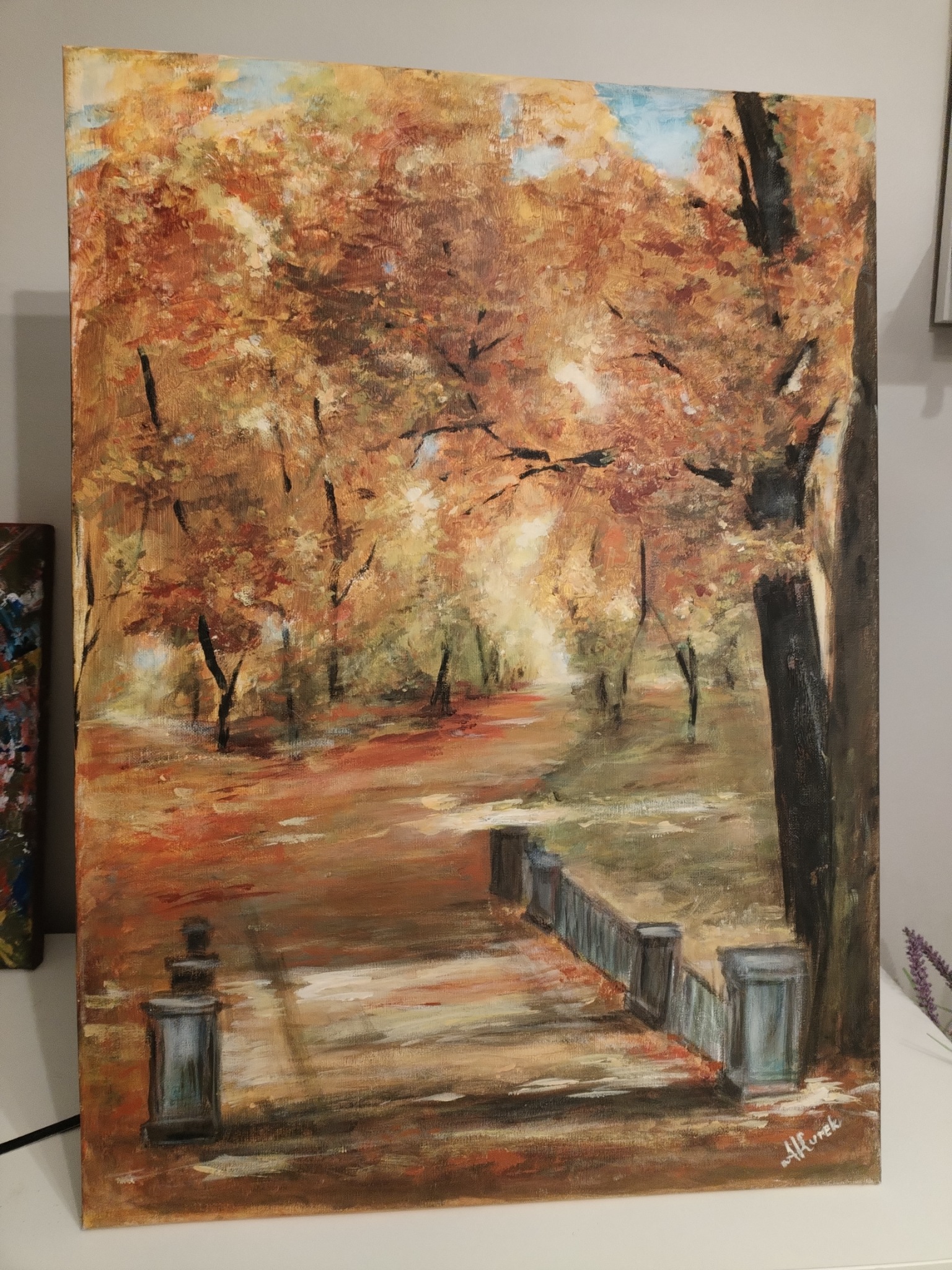 Obraz olejny przedstawiający jesienny park z alejką, drzewami w odcieniach brązu i pomarańczy, oraz fragmentem balustrady. Widoczny podpis autora w prawym dolnym rogu.