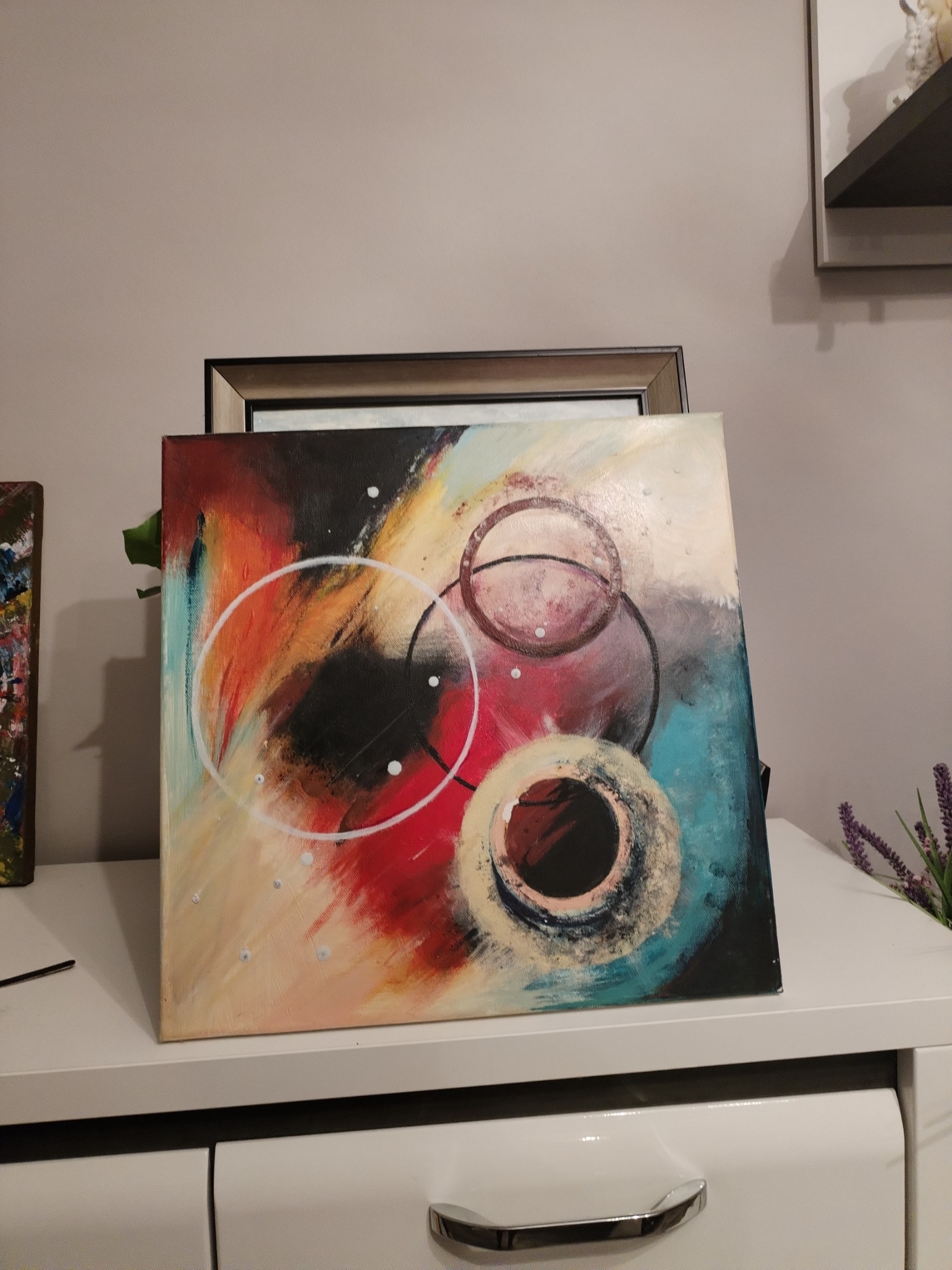 Abstrakcyjne dzieło na płótnie z motywem koncentrycznych okręgów w kolorach czerwieni, błękitu i czerni, umieszczone na białej komodzie, z widocznym fragmentem innego obrazu po lewej stronie...