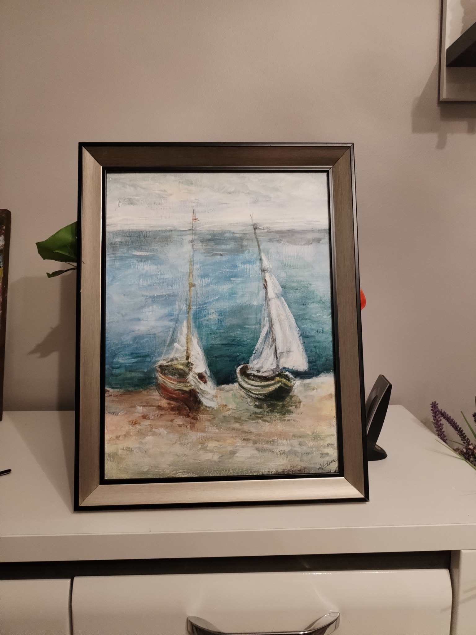 Oprawiony obraz olejny przedstawiający dwa żaglowce na brzegu, w tle błękitne morze i szare niebo, postawiony na białej komodzie.