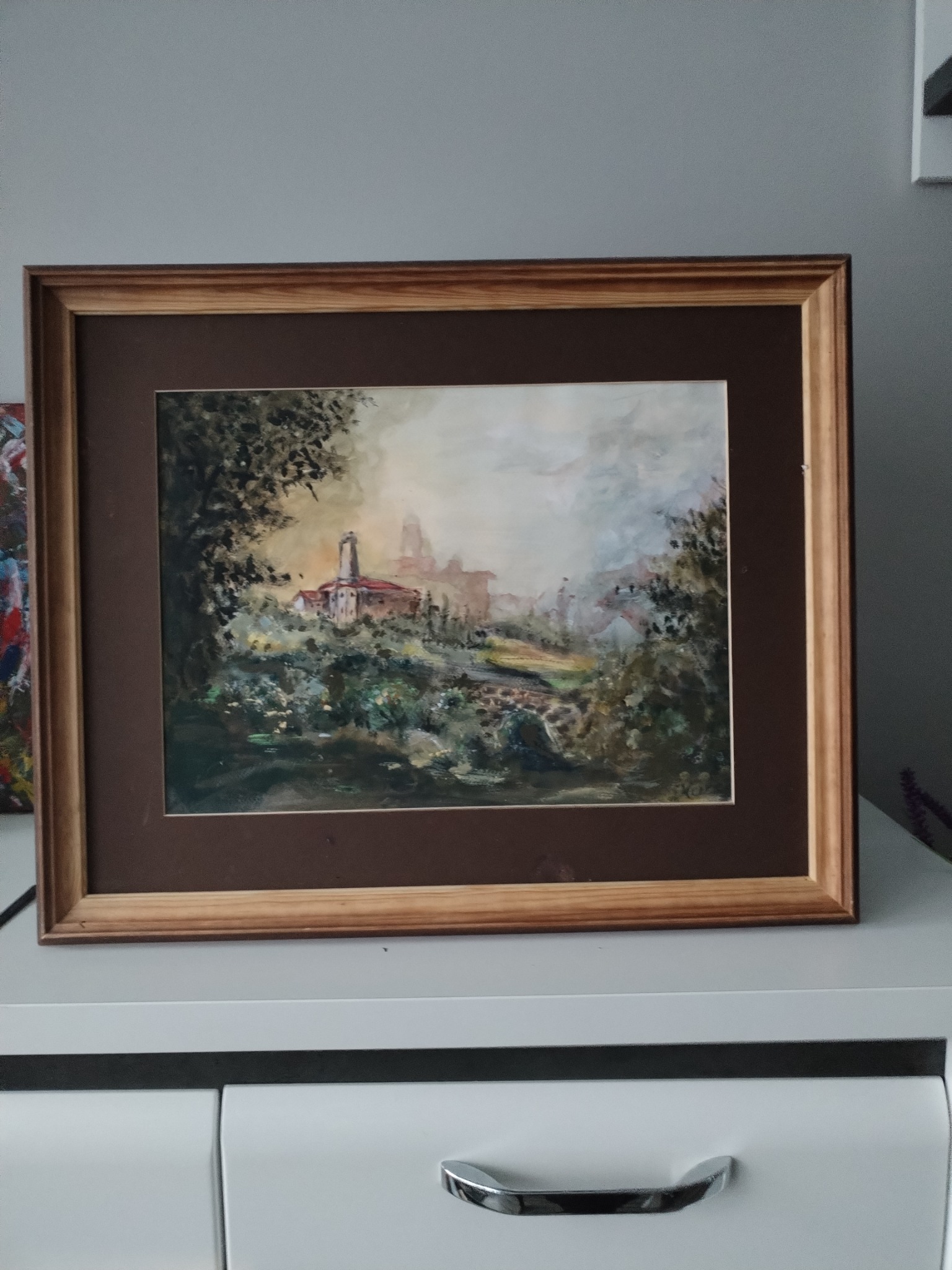 Oprawiony akwarelowy pejzaż przedstawiający toskański krajobraz z wieżą i budynkami w oddali, umieszczony na białej komodzie.