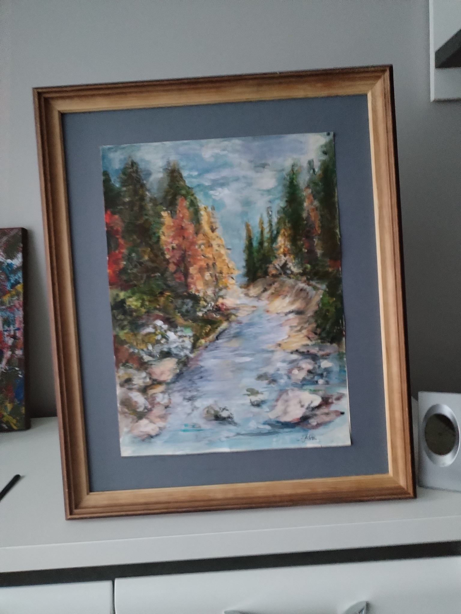 Krajobraz z rzeką i drzewami w jesiennych barwach, namalowany farbami na płótnie, oprawiony w drewnianą ramę z szarym passe-partout, ustawiony na białej komodzie obok głośnika.