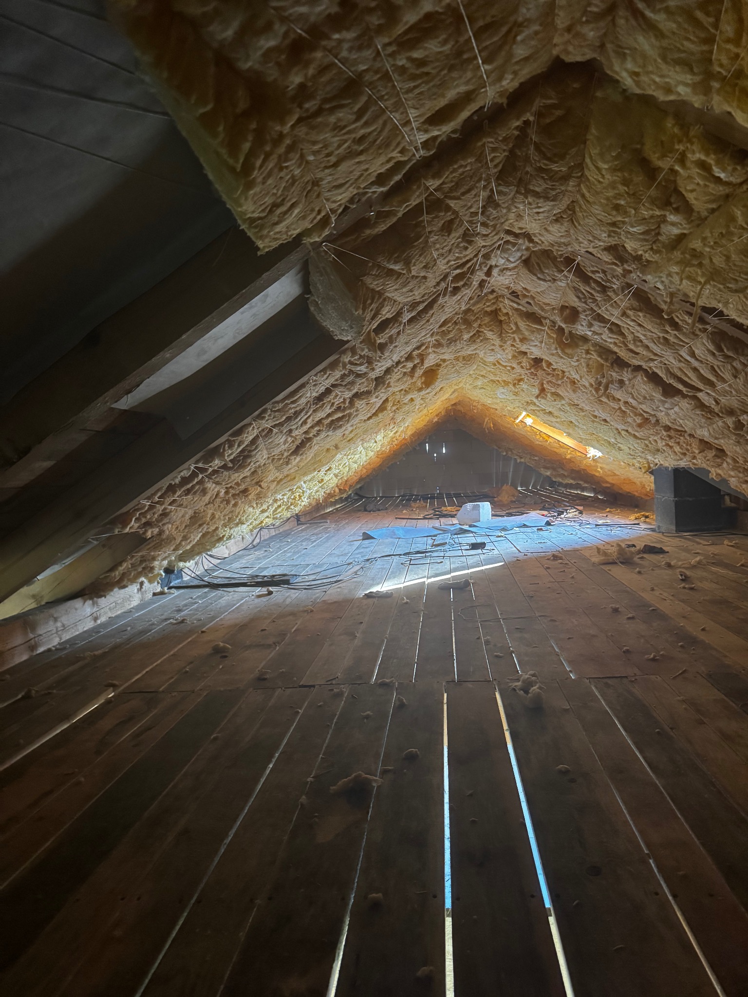 Ocieplone wełną mineralną poddasze z drewnianą podłogą. Widoczne elementy konstrukcyjne dachu i szczeliny w podłodze przepuszczające światło dzienne.