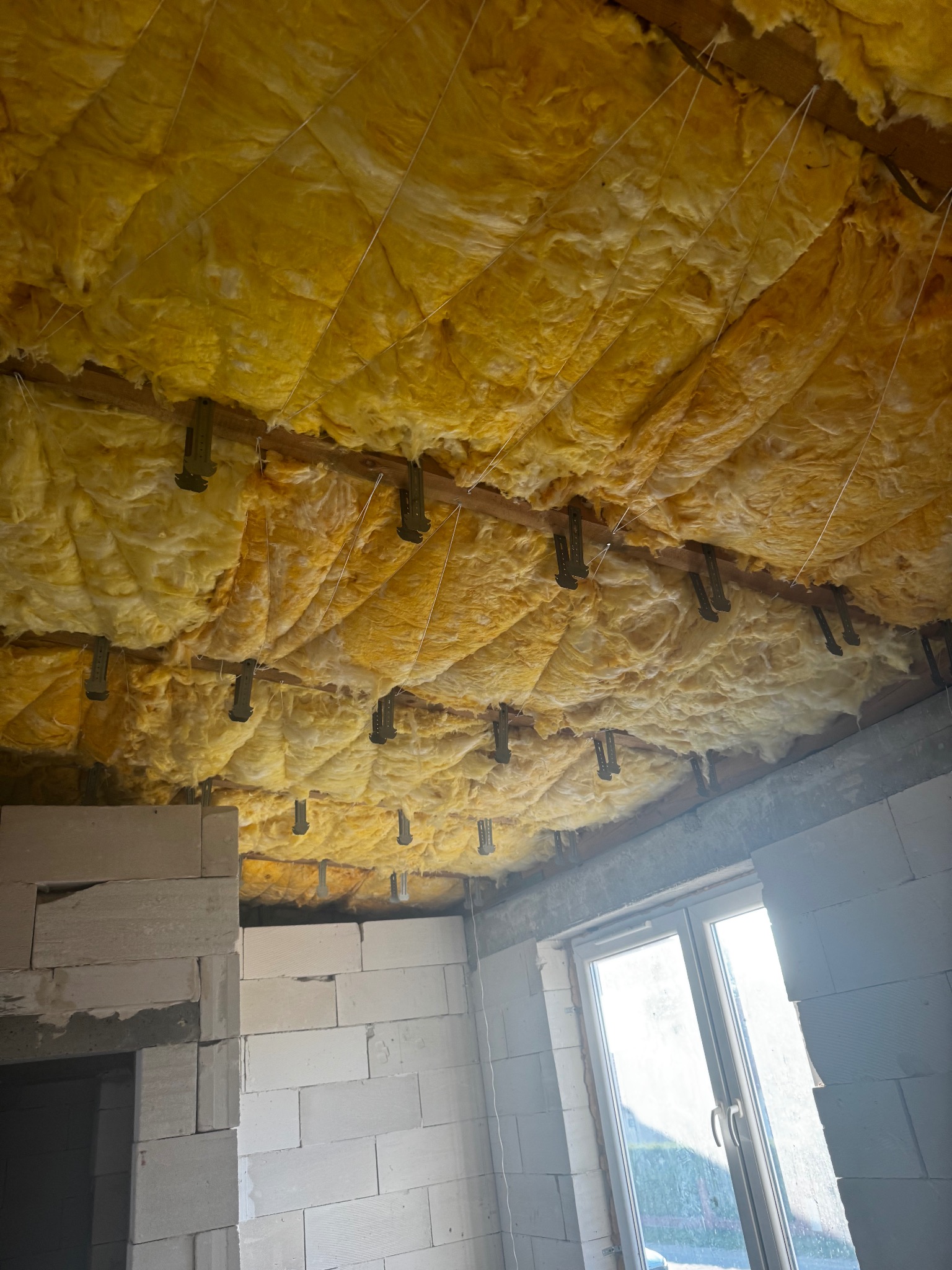 Żółta wełna mineralna zamontowana na poddaszu, widoczne metalowe uchwyty i białe sznurki mocujące. W tle surowa ściana z pustaków i okno.