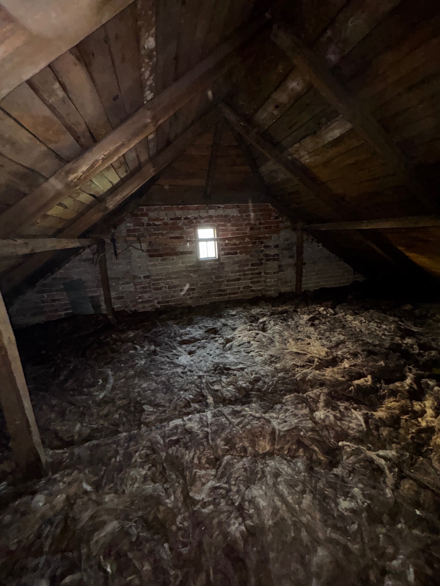 Poddasze z ceglaną ścianą i małym oknem, wypełnione warstwą izolacji termicznej. Widoczne drewniane elementy konstrukcyjne dachu.