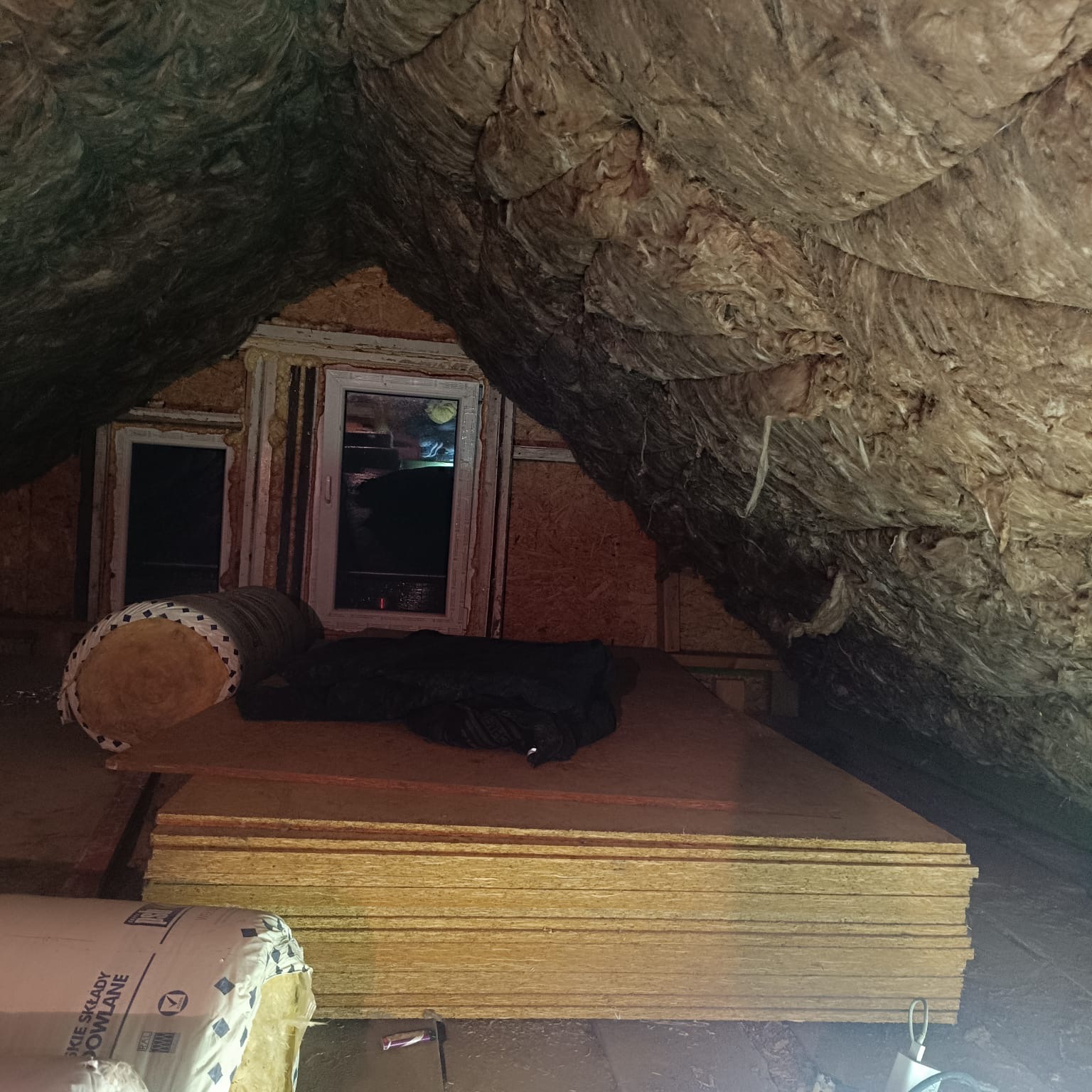 Poddasze z oknami, częściowo ocieplone wełną mineralną, z widocznymi płytami OSB i rolką izolacji. Prace w toku, widoczne materiały budowlane.