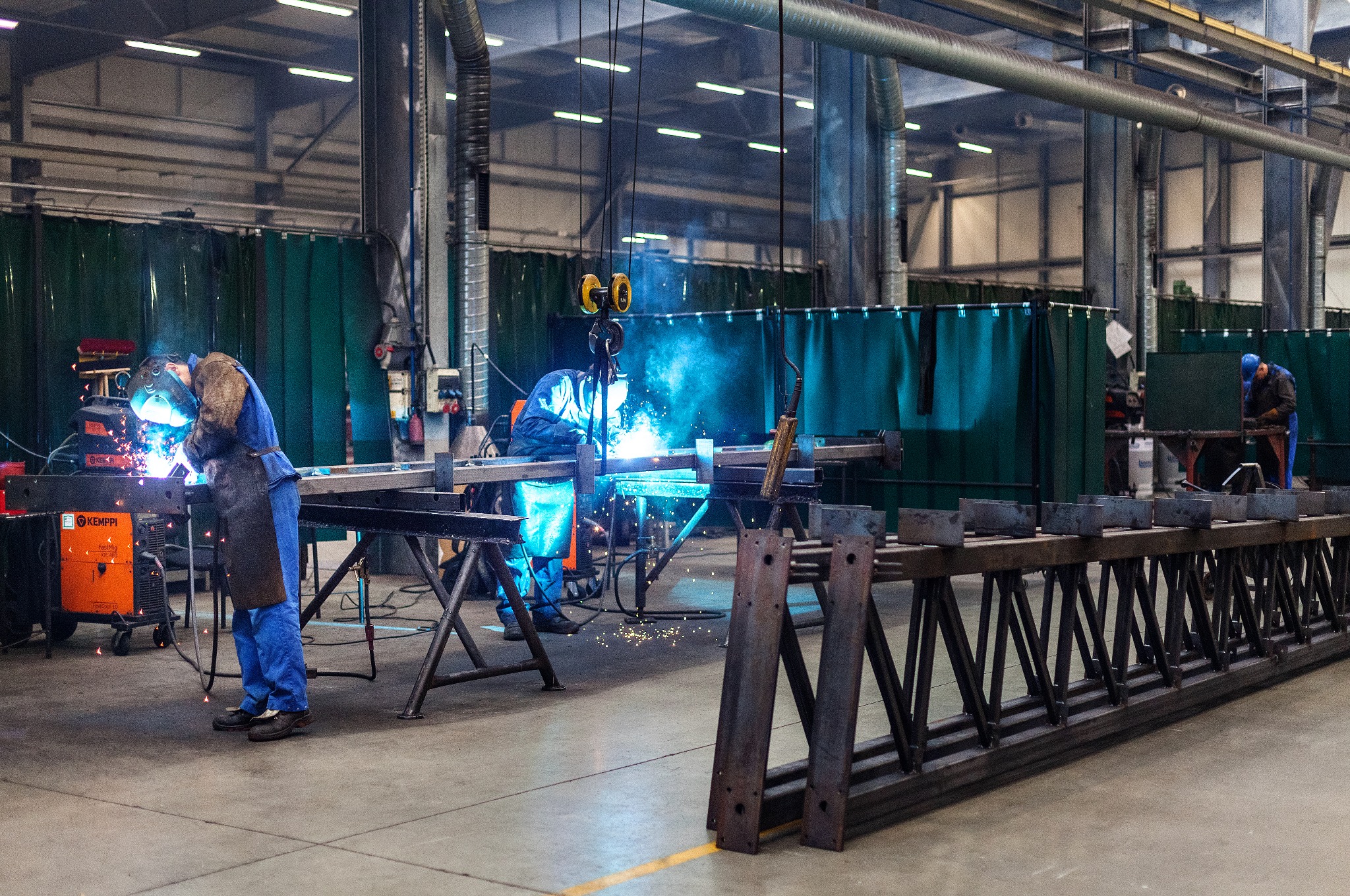 Spawanie konstrukcji stalowej w hali produkcyjnej, widoczne iskry i spawacze w kombinezonach ochronnych, dźwig nad głową, w tle zielone zasłony spawalnicze.