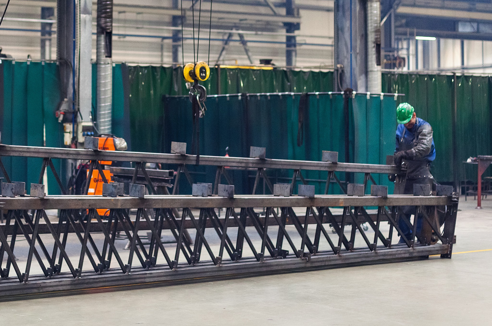 Pracownik w kasku obrabia metalową konstrukcję kratownicową w hali produkcyjnej, widoczny suwnicowy hak i osłony spawalnicze.