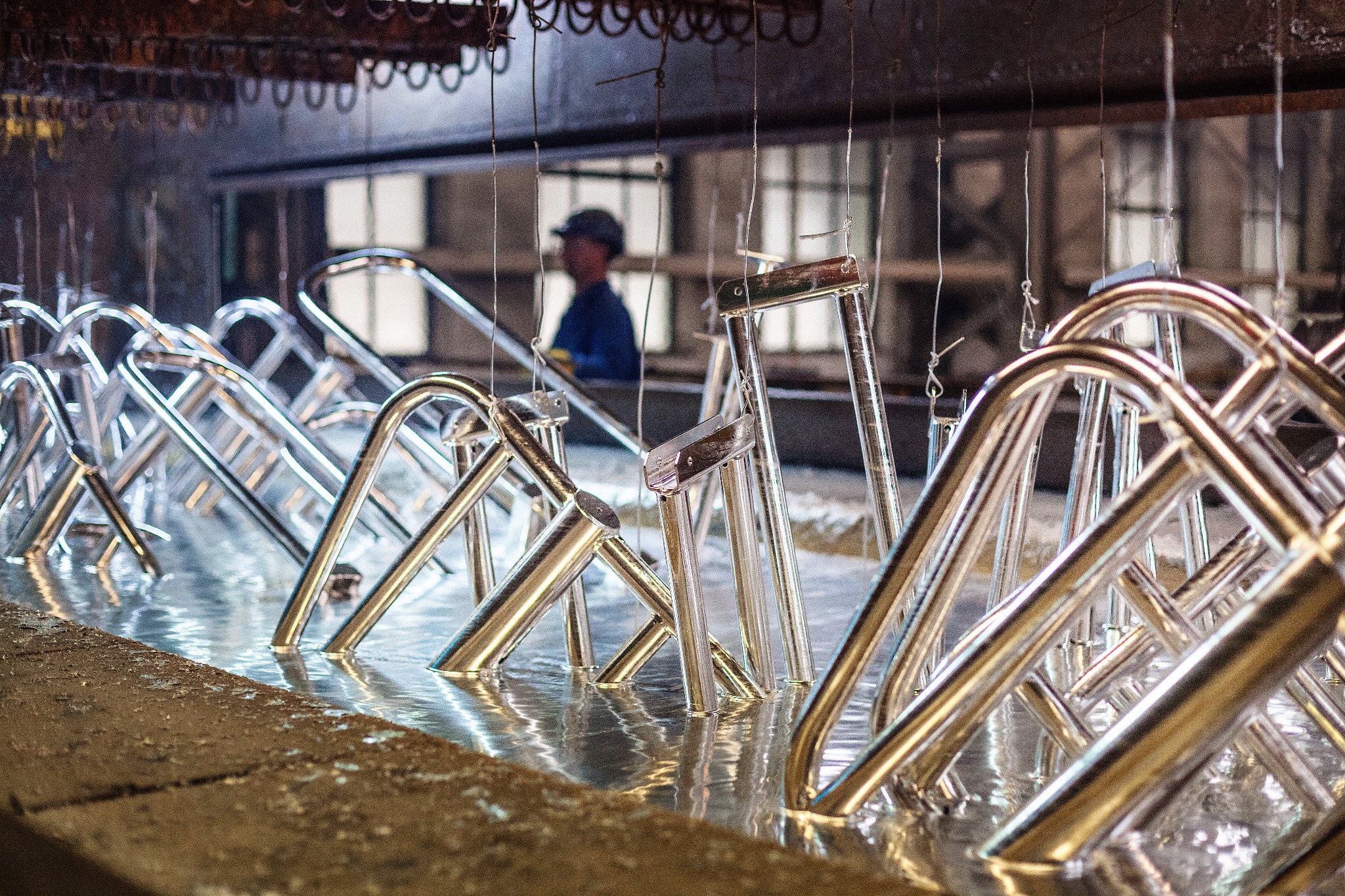 Metalowe elementy konstrukcyjne zanurzone w roztworze podczas procesu obróbki, widoczny pracownik w tle.