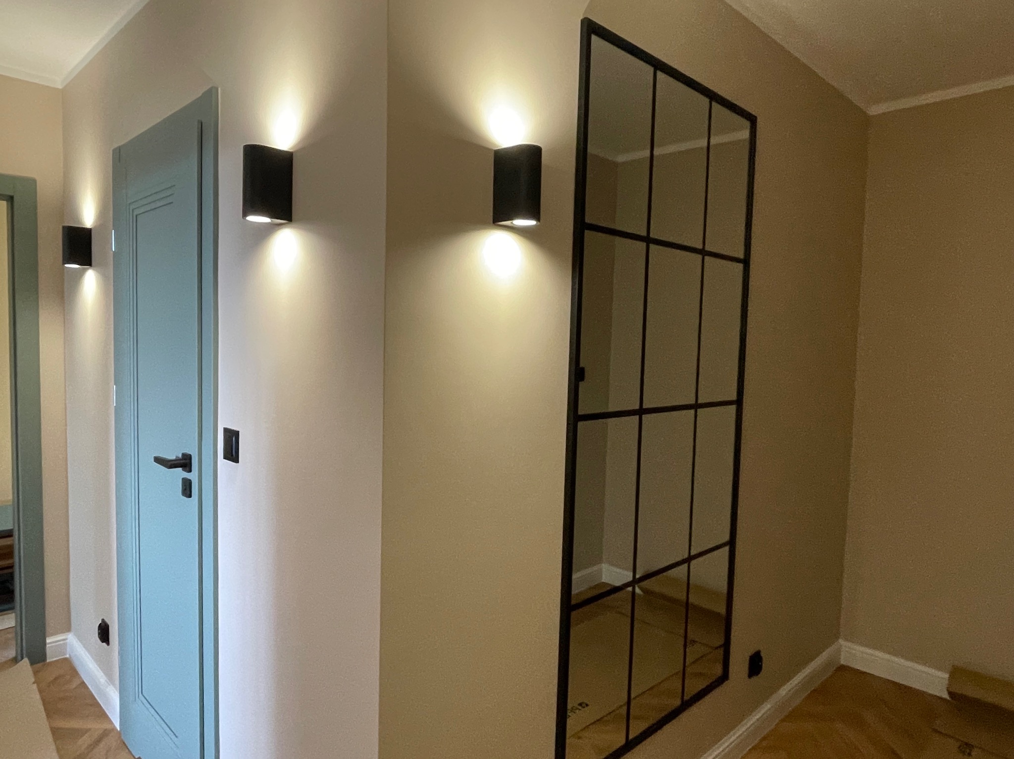 Korytarz po remoncie z pomalowanymi ścianami w beżowym odcieniu, błękitne drzwi z czarną klamką, czarne, nowoczesne kinkiety ścienne emitujące ciepłe światło i czarna ścianka działowa ze szklanymi...