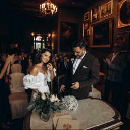 AGENCJA REKLAMOWO-FOTOGRAFICZNA MARCIN TORBIŃSKI - Para młoda w eleganckim wnętrzu, otoczona gośćmi podczas ceremonii zaślubin. W tle obrazy w złotych ramach i żyrandol.