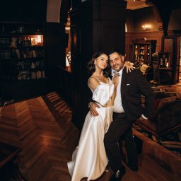 AGENCJA REKLAMOWO-FOTOGRAFICZNA MARCIN TORBIŃSKI - Elegancka para w objęciach na tle wnętrza w stylu retro. Kobieta w białej sukni, mężczyzna w ciemnym garniturze, parkiet w jodełkę, biblioteczka w tle.